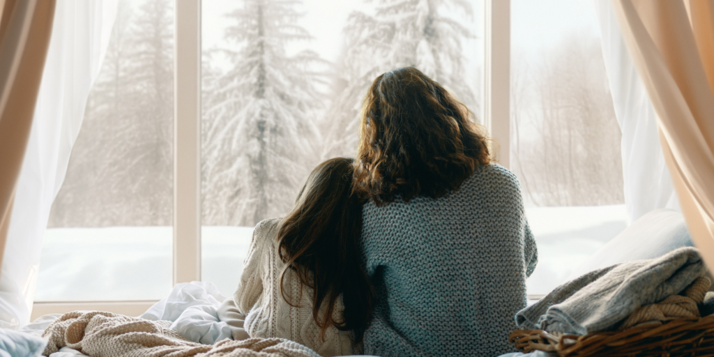 Mother And Daughter Looking Out Snowy Window In Winter - The Best Time of Year to Buy a New Furnace
